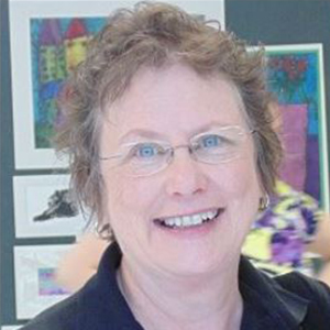 Barbara Laws wears thin rimmed glasses and smiles looking at the camera. There is artwork on walls in the background.
