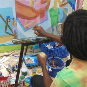 Mensah is painting a colorful picture at his easel. His back is turned and you see his head and arm in motion.