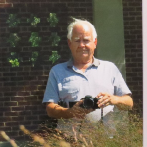 Gray haired male holds a camera in a blue shirt and looks directly at the photographer