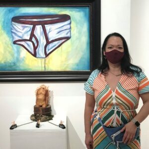 Samantha Wegener at the Offsite Gallery standing wearing a maroon mask in front of her painting of Dad Bod and her sculpture of a male torso