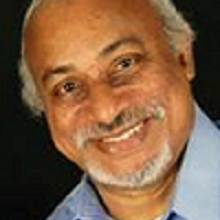Dark skinned gentlemen with gray hair and moutache and short beard slants head to the right as he smiles directly looking at the camera