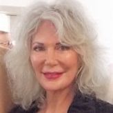 Lady with shoulder length wavy gray hair half grinning and staring directly at the camera. She has bright pink lipstick.