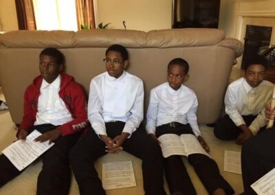 4 young boys sit on a couch in black pants and white shirts inside a house