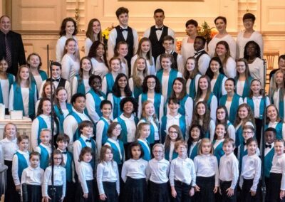 Approx. 30 male and female young singers stand together wearing bright blue vests and white shirts. The directors surround them wearing black.